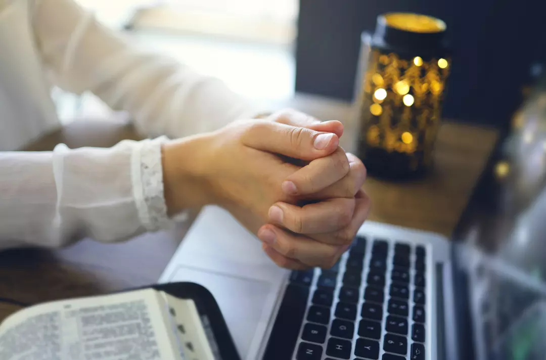 Someone praying while using their laptop with their Bible open next to them