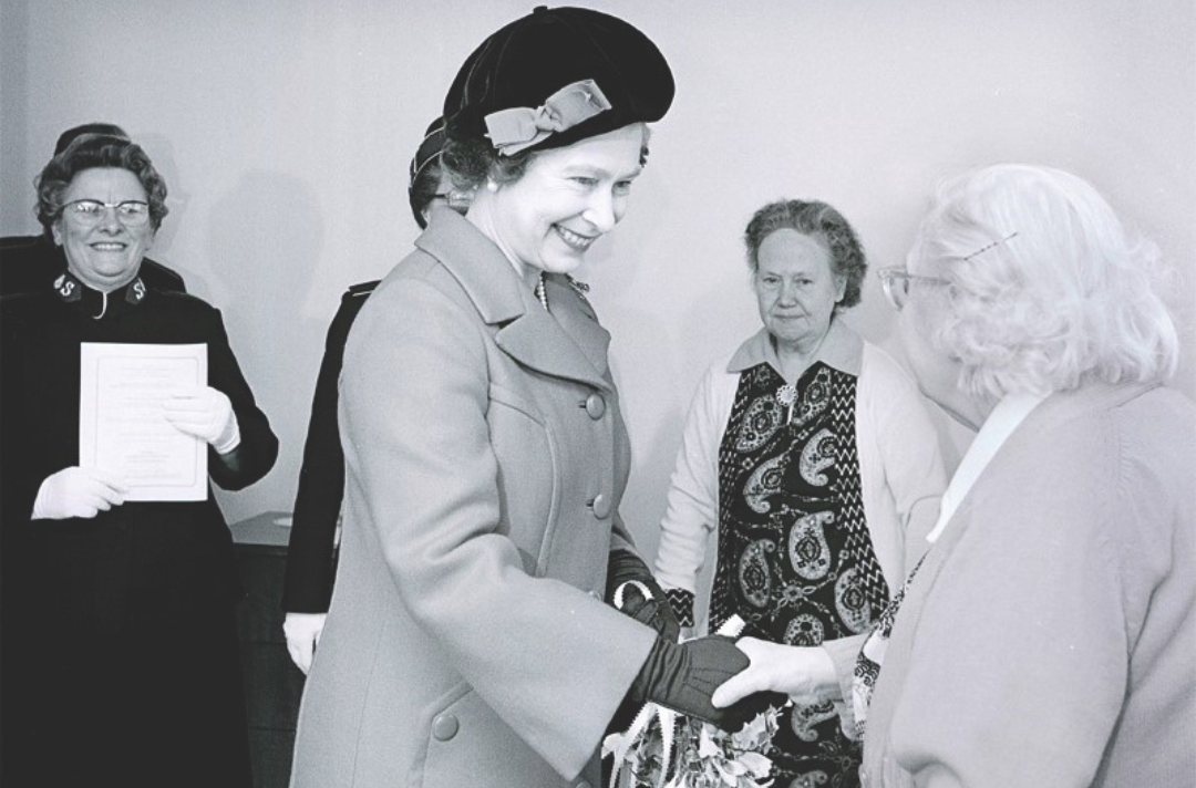 The Queen Elizabeth II shaking hands of an older woman with a Salvationist in uniform looking on