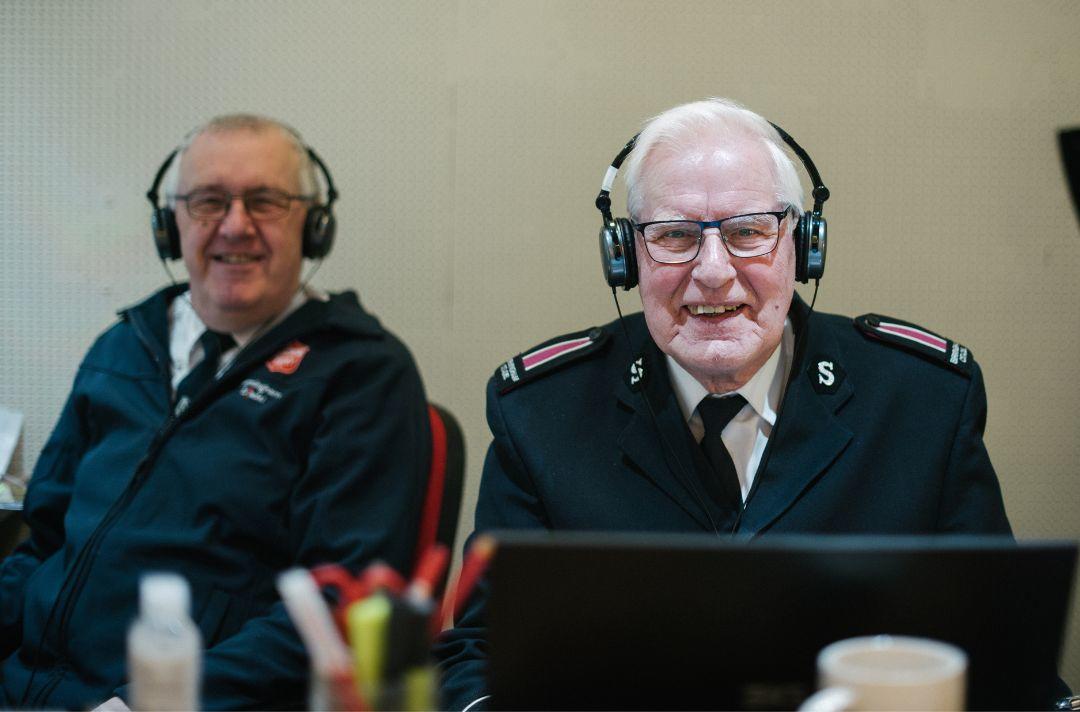 Two older people wearing Salvation Army uniform with headphones sitting at an AV desk