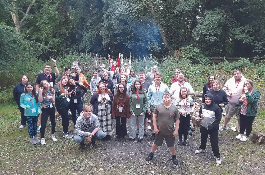 A group of teenagers and adults in a woodland clearing by a camp fire