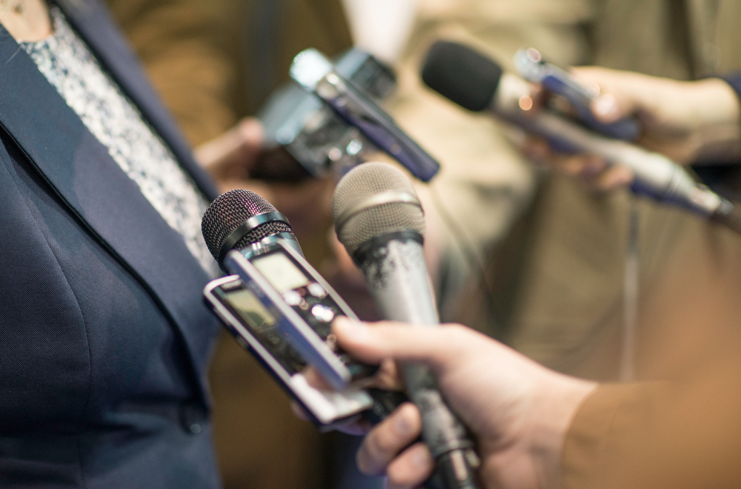 Someone wearing smart clothes being interviewed with microphones and recording devices surrounding them