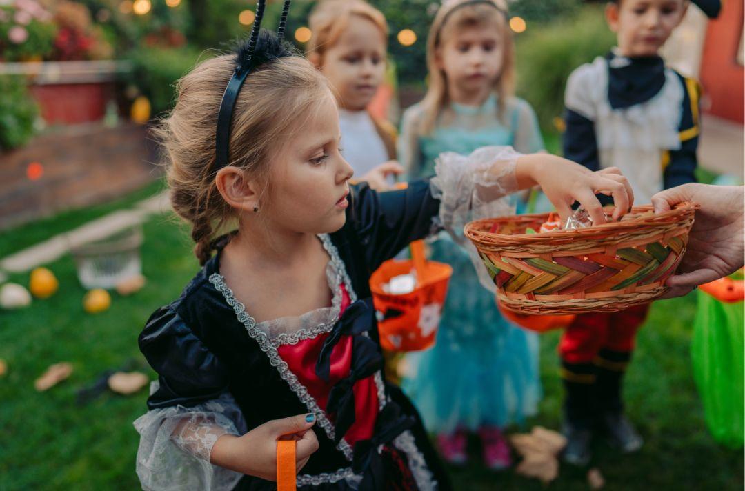 A photo of children trick-or-treating. The children are wearing fancy dress outfits - one is a princess - and they are picking sweets out of a basket