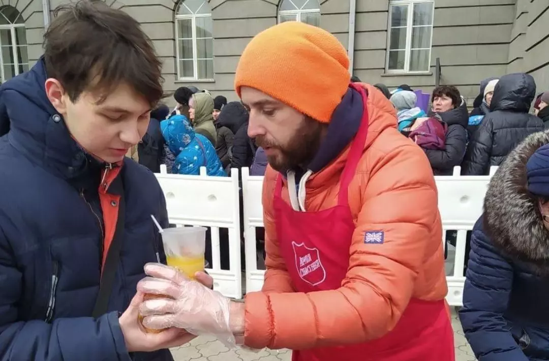 A man wearing a Salvation Army apron giving someone a drink. They are standing in front of a queue of people. Everyone is wrapped up in coats and hats.