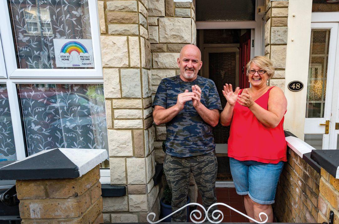 A photo of a couple standing outside their front door clapping and smiling with a rainbow sign in their front window