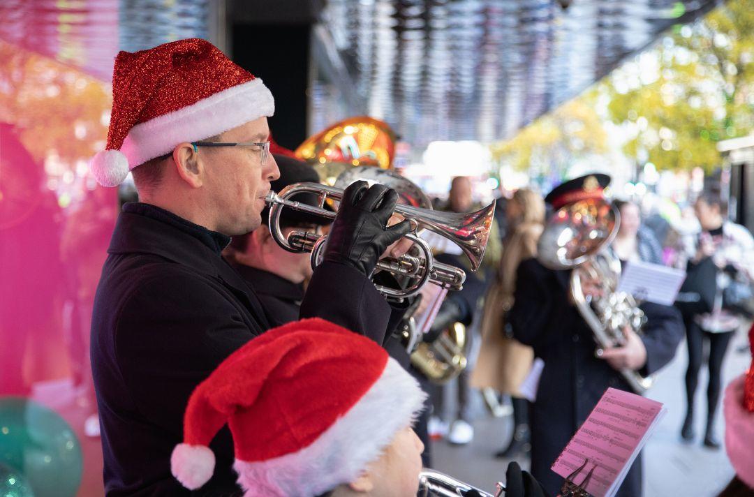 A photo of a Salvation Army brass band carolling on a high street. The musicians are adults and children and they are wearing Salvation Army uniform and Father Christmas hats.