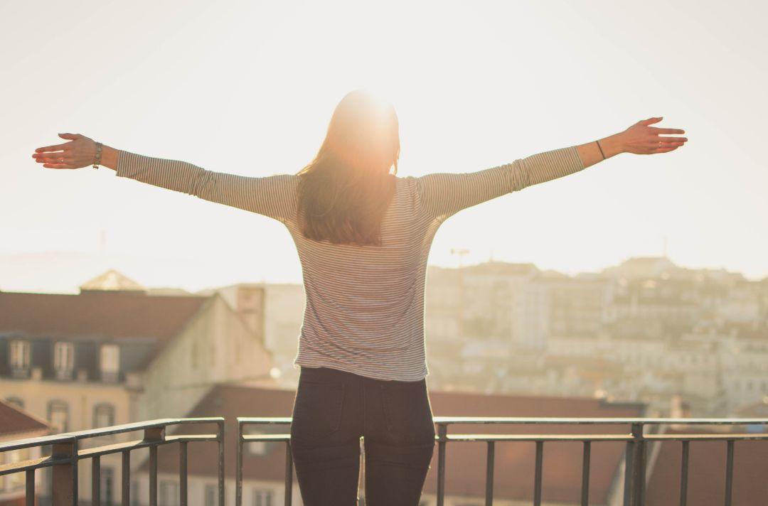 A photo of someone standing on a balcony in a city with their arms outstretched facing the sunshine