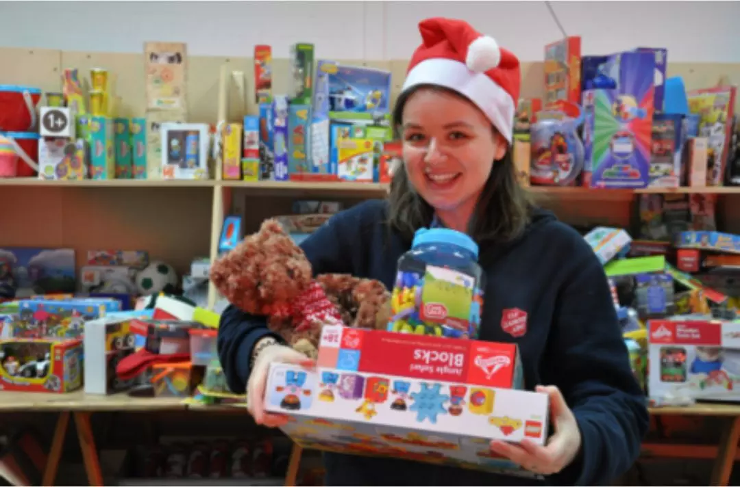 A photo of a Salvation Army volunteer wearing a Santa hat and holding a pile of donated toys in a warehouse full of toys