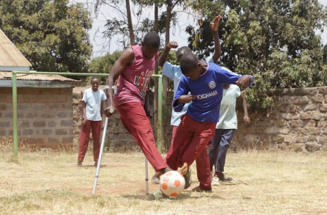 A photo of two black boys playing football outside on dry grass. One boy is using crutches.