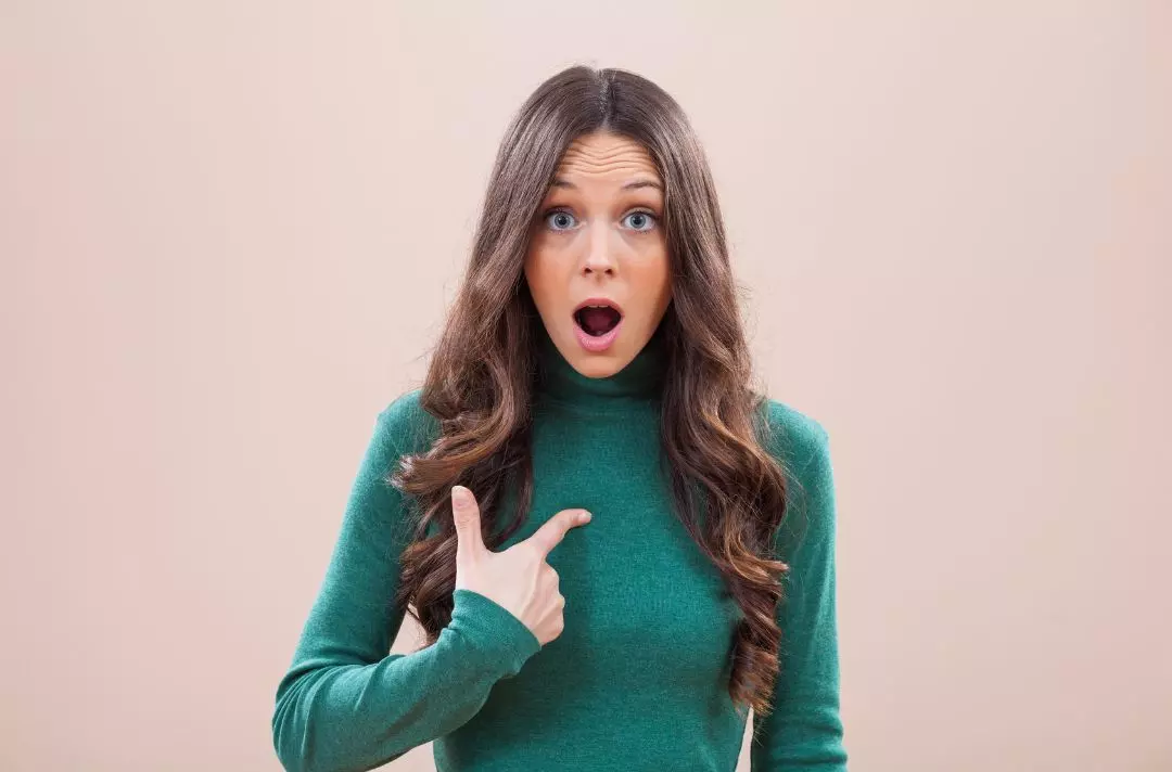 A photo of a woman pointing to herself with her mouth open, as if she is saying 'what, me?'