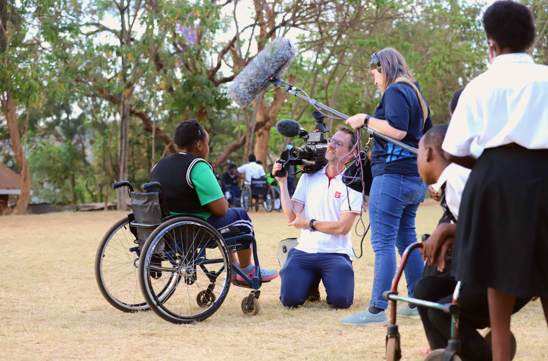 Image shows behind the scenes of a self-denial video. A young person in a wheelchair talks to a camera while other young people stand around and watch.