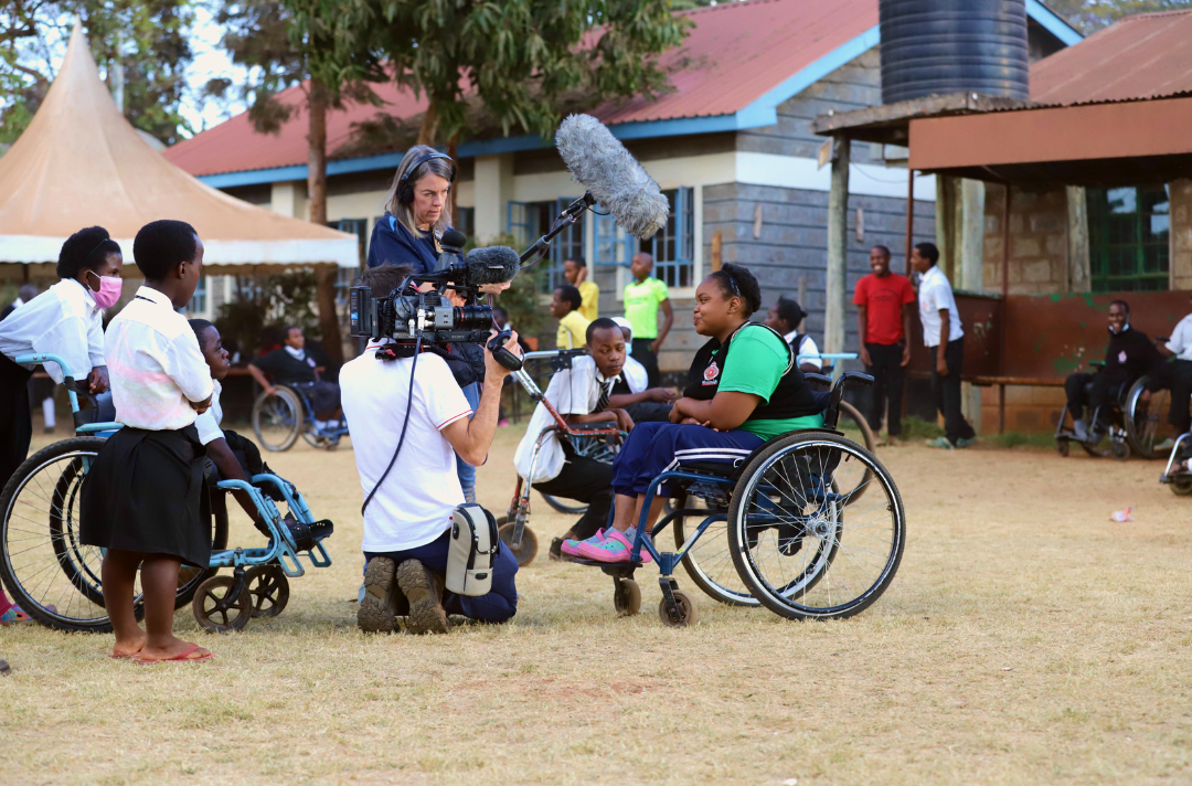 Image shows behind the scenes of a self-denial video. A young person in a wheelchair talks to a camera while other young people stand around and watch.