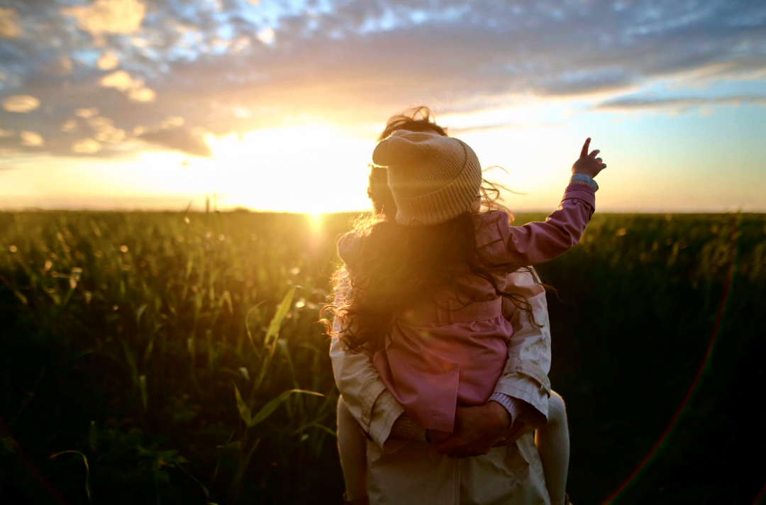 Photo shows a child being held by an adult in a field filled with tall plants while the sun is low in the sky.