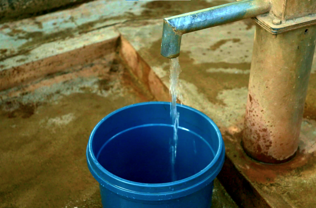 Photo shows a blue bucket being filled with water from a pump.