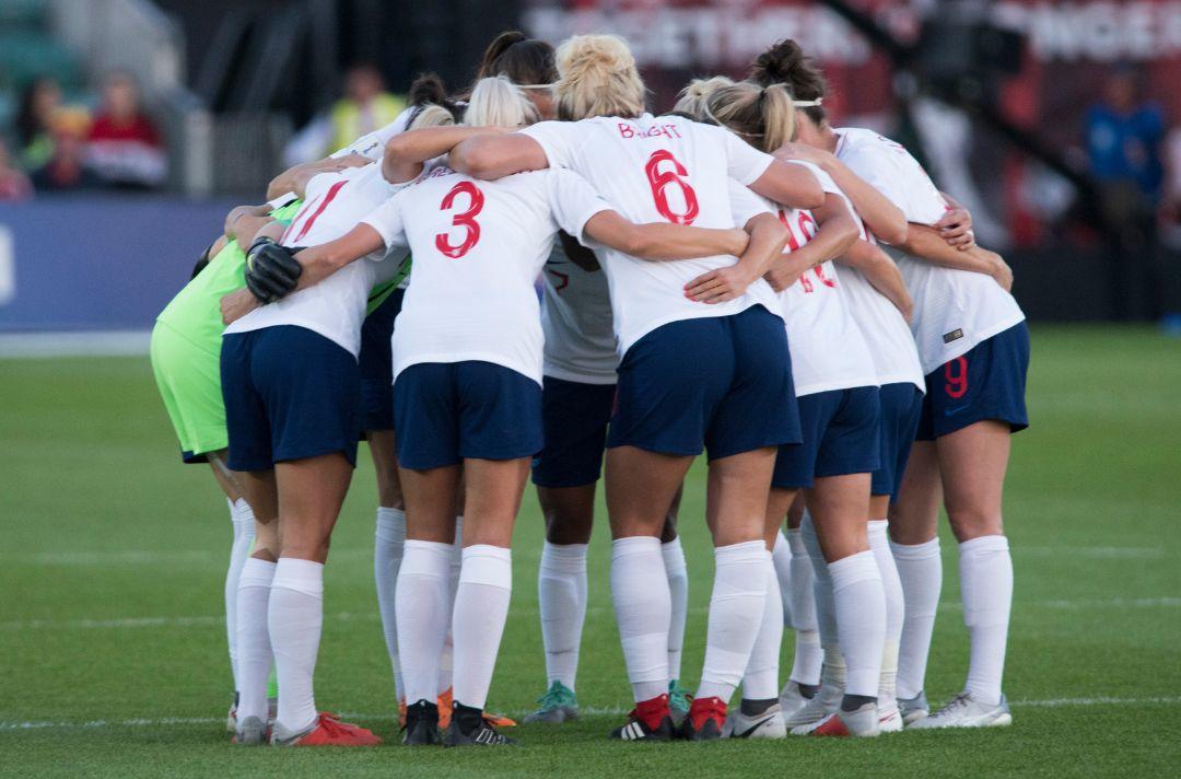 A photo of the England women's football team in a group huddle on a football pitch