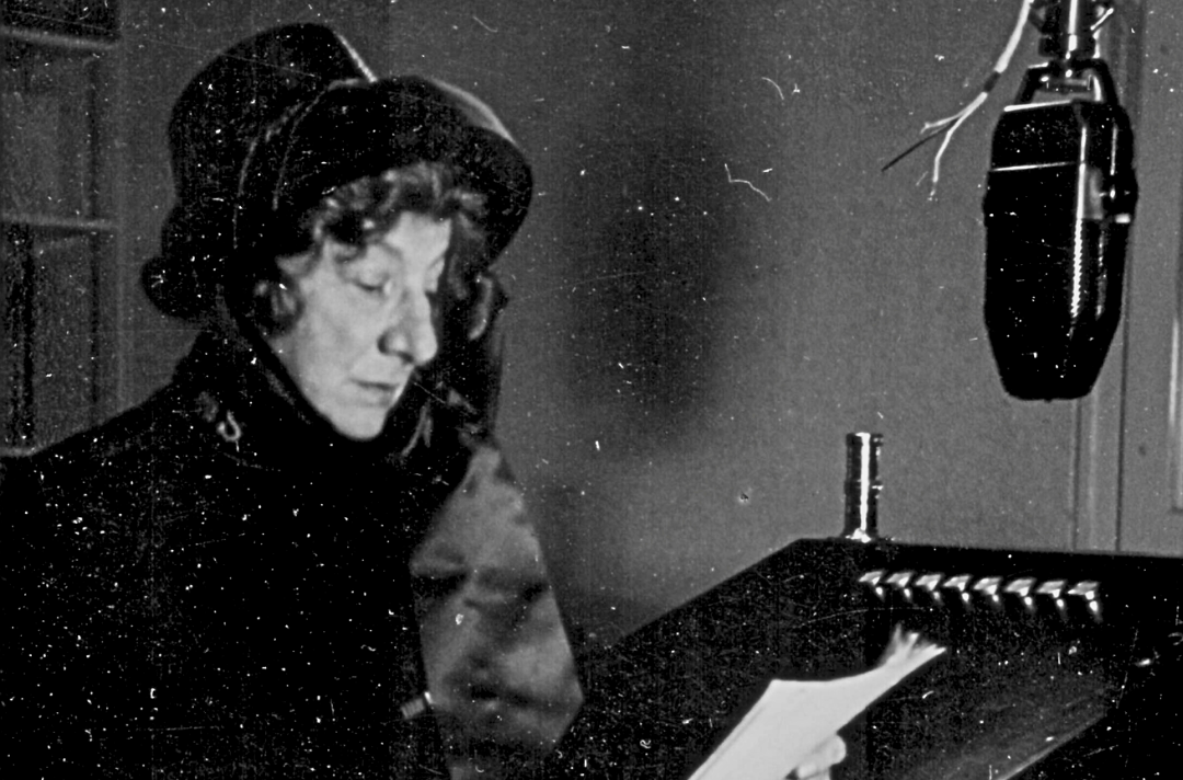 A black and white photo shows Evangeline Booth reading from a piece of paper as she speaking into a microphone.