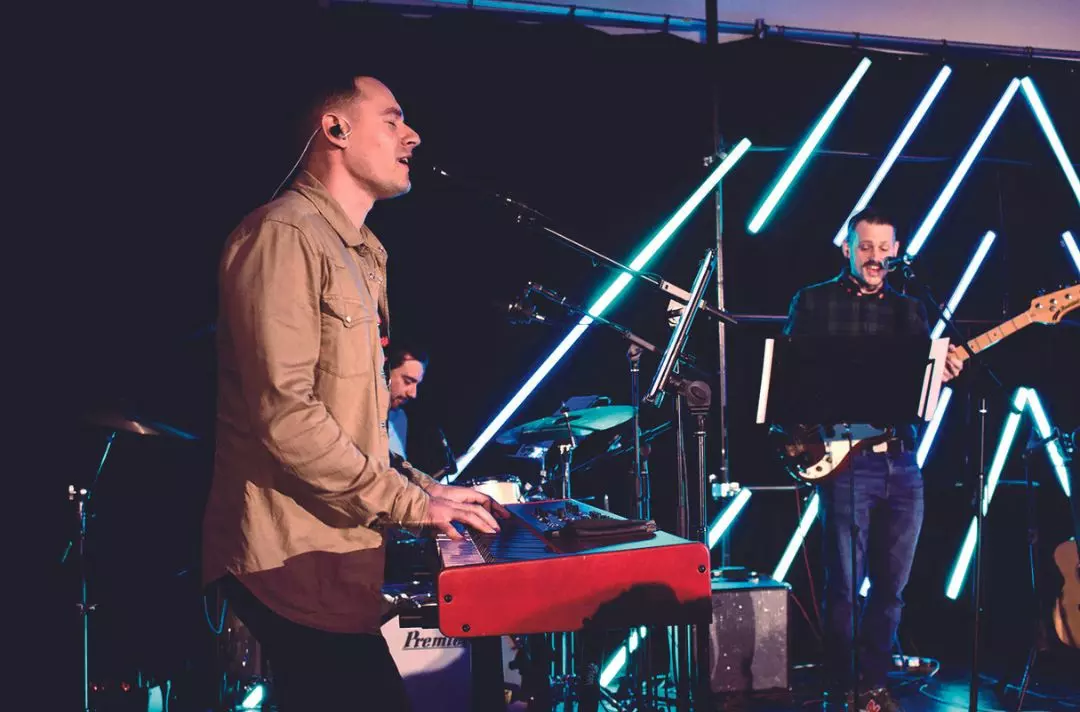 A photo of worship at Shalom, with someone singing and playing a keyboard, someone playing drums and someone singing and playing the guitar