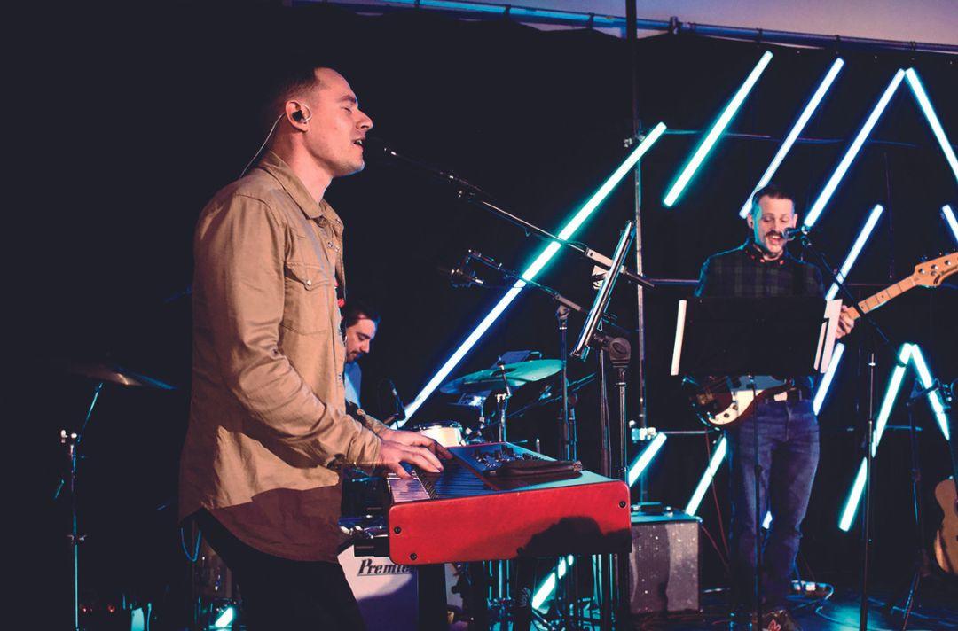 A photo of worship at Shalom, with someone singing and playing a keyboard, someone playing drums and someone singing and playing the guitar