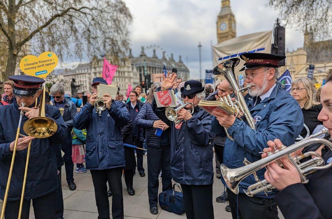 A photo of a Salvation Army brass band playing in Parliament Square, surrounded by a crowd hold placards. Big Ben is visible in the background.