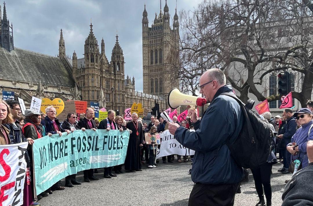 A photo of Commissioner Anthony Cotterill praying into a megaphone in front of a crowd hold placards in Parliament Sqaure.