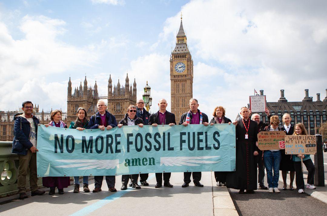 A group photo on Westminster Bridge with Big Ben in the background. People are holding a banner that reads 'No more fossil fuels. Amen.' In the group is Commissioner Anthony Cotterill, Cadet Lizzy Kitchenside and a child holding a placard with a Salvation Army red shield on it.