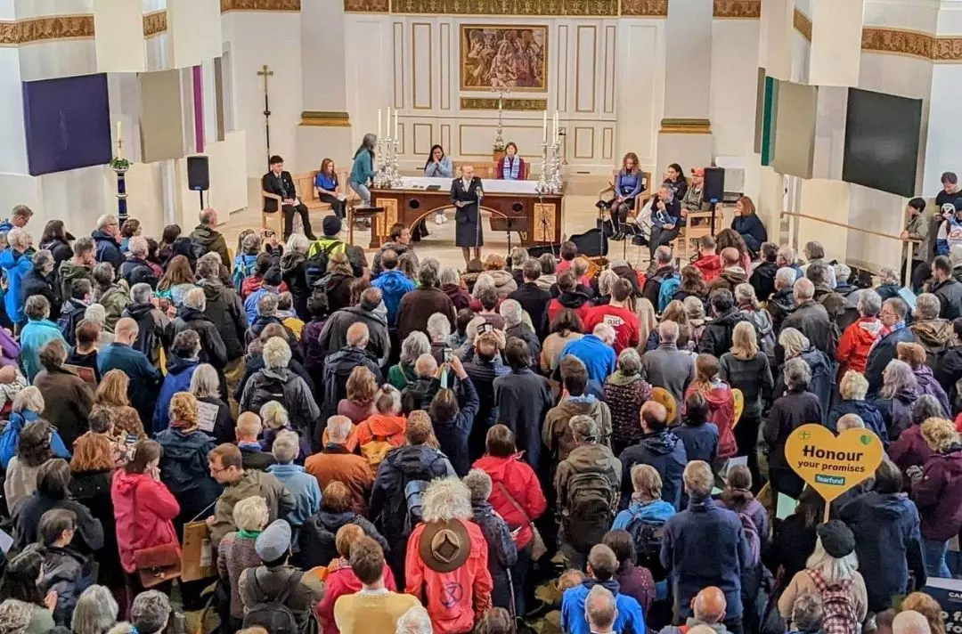 A photo of a congregation standing in the body of a church listening to Cadet Lizzy Kitchenside speak from the front into a microphone