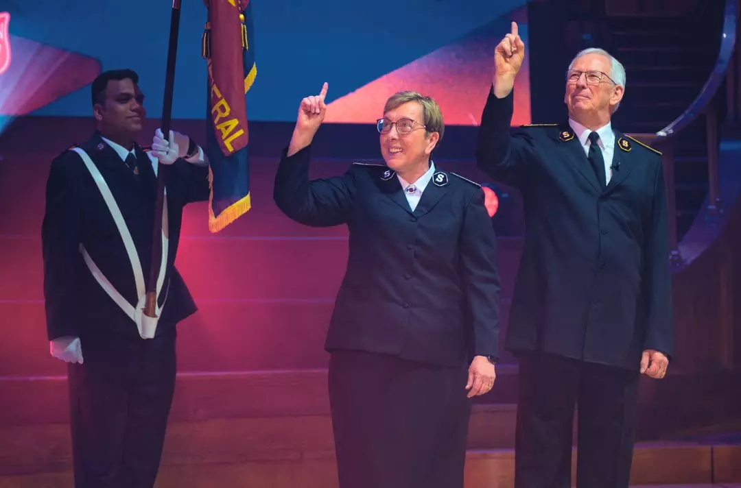 A photo of General Brian Peddle and Commissioner Rosalie Peddle on stage in Salvation Army uniform giving The Salvation Army salute - an index finger raised to Heaven. there is a Salvation Army officer holding a Salvation Army flag in the background.
