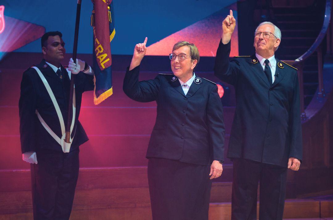 A photo of General Brian Peddle and Commissioner Rosalie Peddle on stage in Salvation Army uniform giving The Salvation Army salute - an index finger raised to Heaven. there is a Salvation Army officer holding a Salvation Army flag in the background.