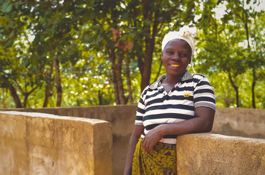 Photo shows a woman smiling in front of a low structure and trees.