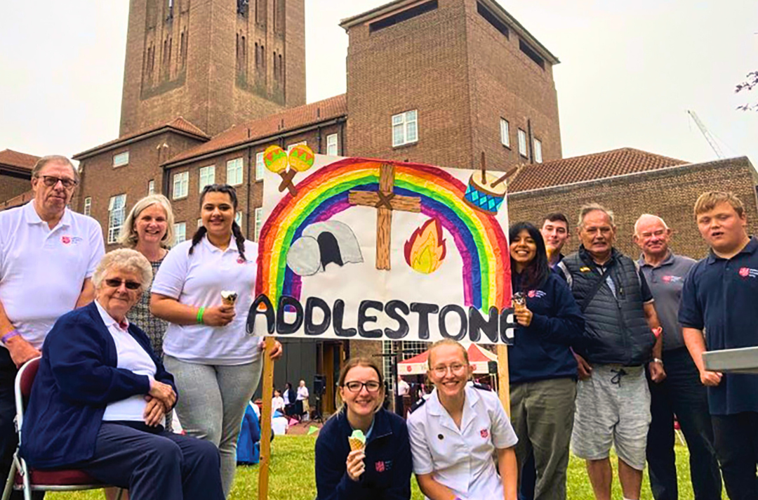 Photo shows people connected to the Addlestone Corps gap year holding a sign that reads: 'Addlestone' and is surrounded by music instruments, a tomb, a flam, a cross and a rainbow.