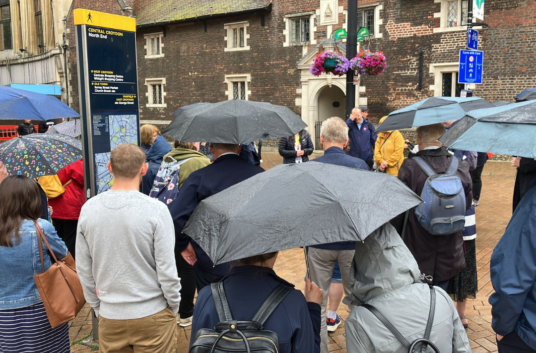 A photo of a large group gathering outside an old building in Croydon, praying in the rain. People are standing under umbrellas. 