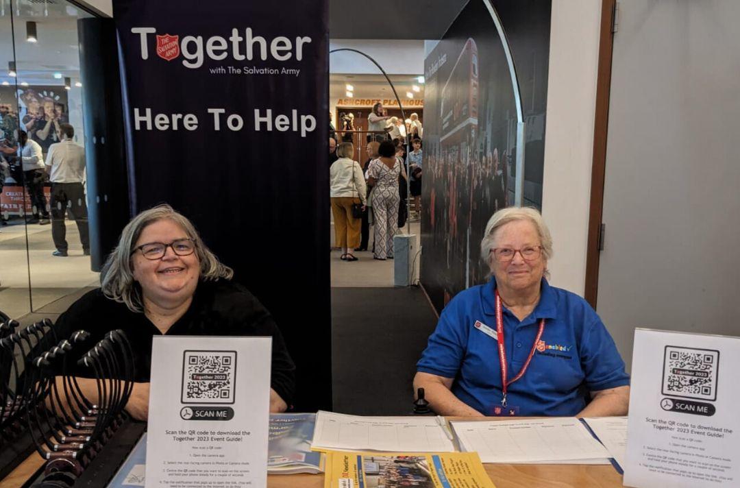 A photo of two woman sitting at a welcome desk with a sign that reads 'Together 2023: Here to help'
