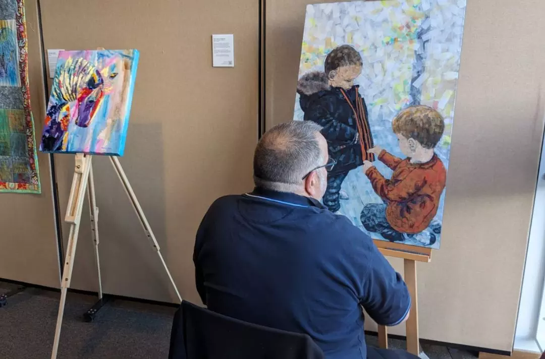 A photo of a man painting a picture of two children helping one another on an easel