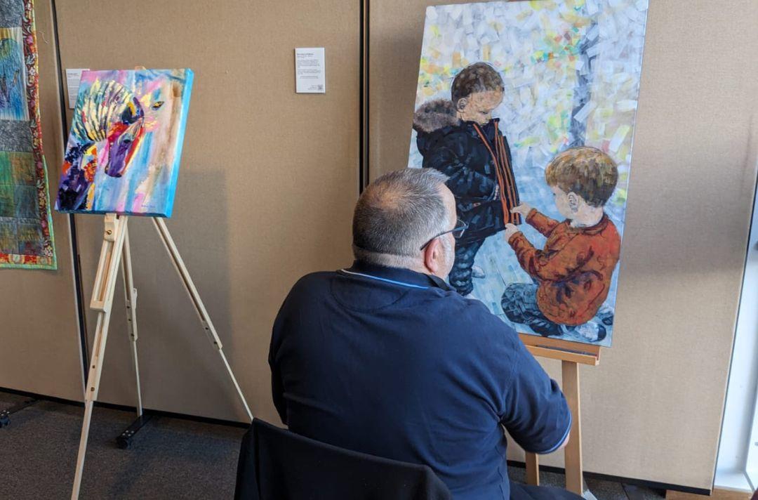 A photo of a man painting a picture of two children helping one another on an easel 