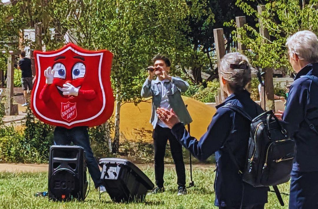 A photo of Charlie Green singing in a park next to a person wearing a Salvation Army red shield costume