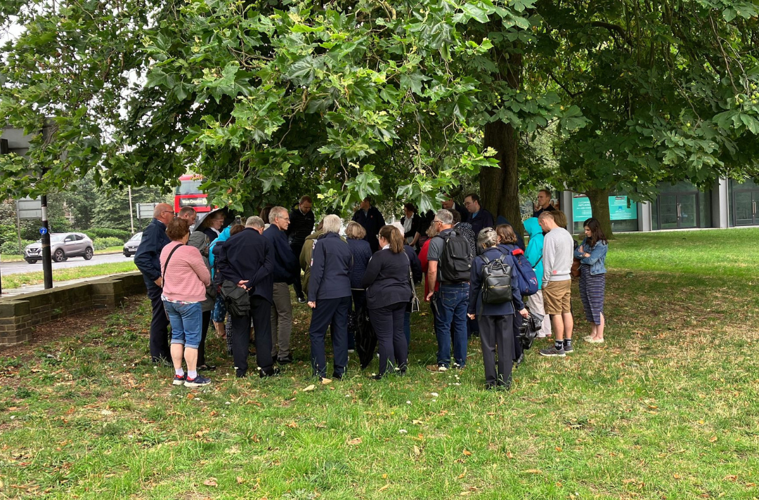 A photo of a group gathered under a tree praying, taking shelter from the rain. A London bus is driving down the road next to the tree.