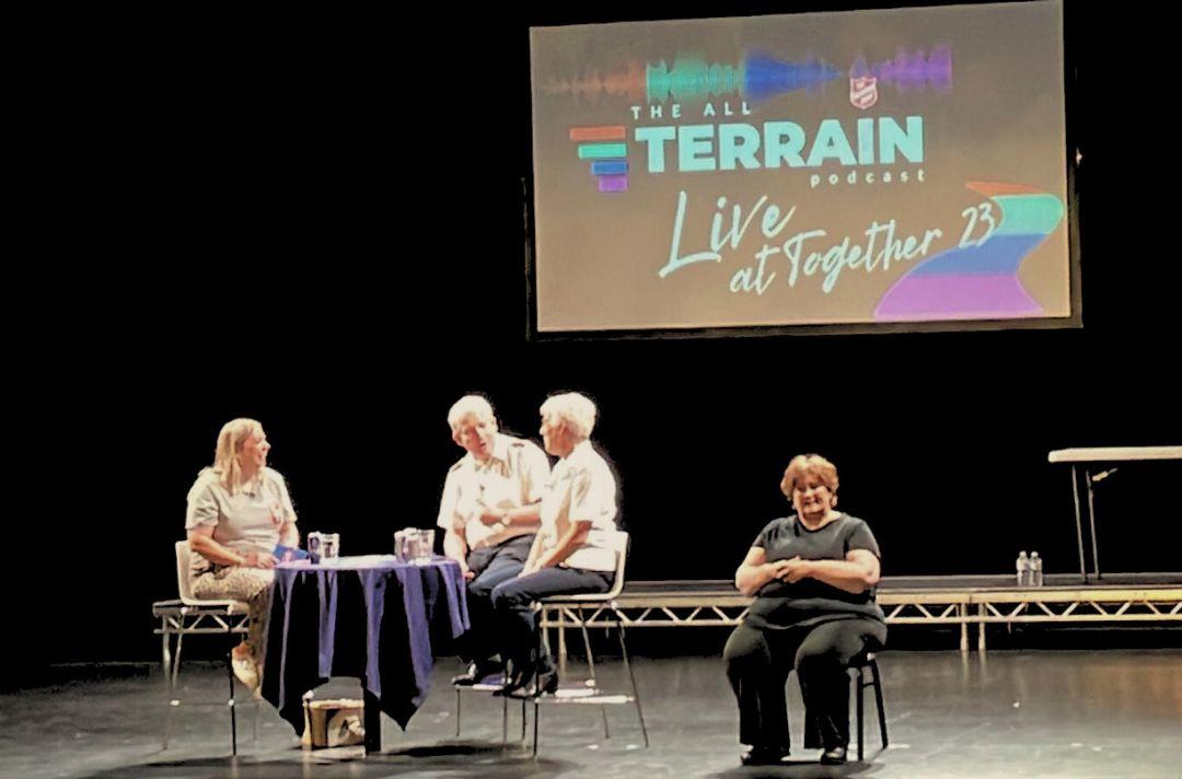A photo of Jo Taylor speaking to Paul and Jenine Main. They are seated on a stage with a BSL translator sitting next to them.