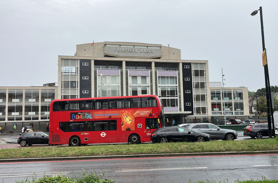 A photo of Fairfield Halls, a large concert hall. The photo is taken on a rainy day, looking at the building from the other side of a busy road with cards and a red London bus.
