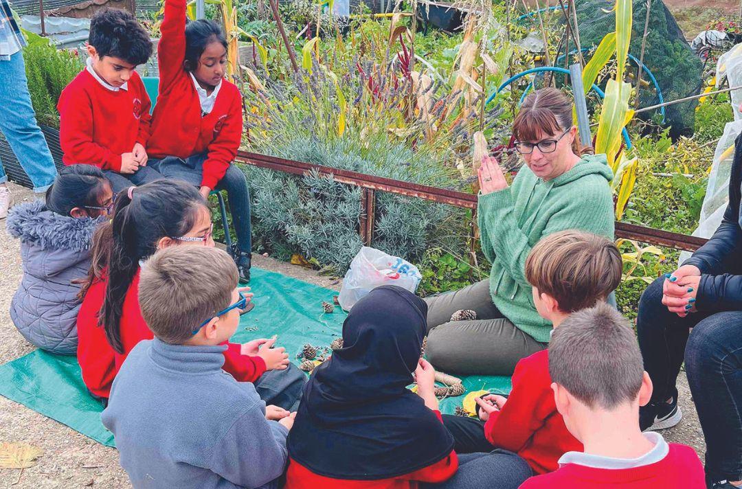 A photo of an allotment with school children sitting and standing around a lady talking.