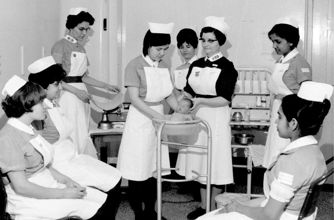 A black and white photo of a group of nurses standing and sitting around two nurses bathing a baby in a small tub