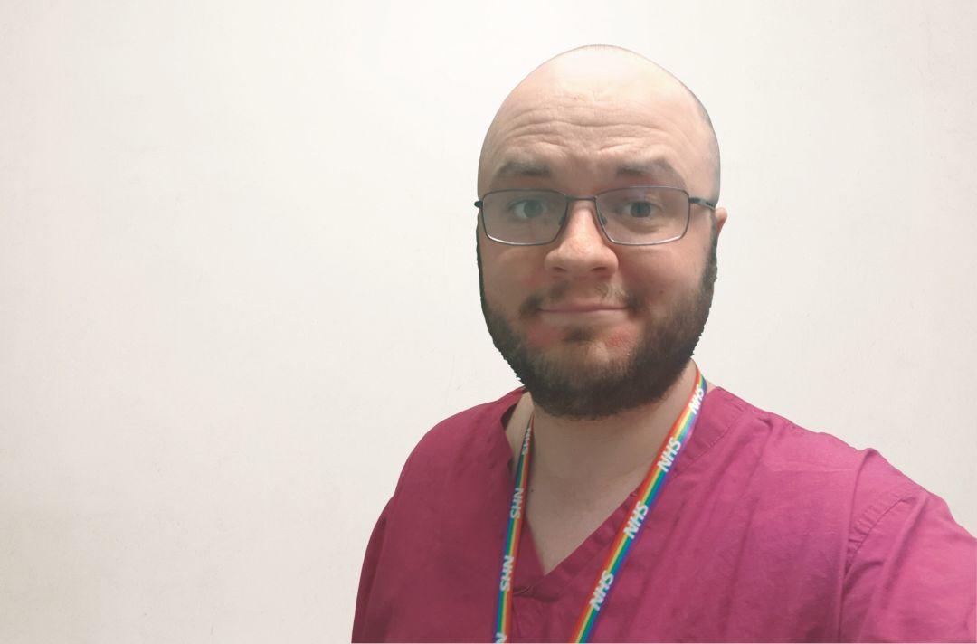 A photo of Dan Holland in nurses scrubs, wearing a rainbow NHS lanyard