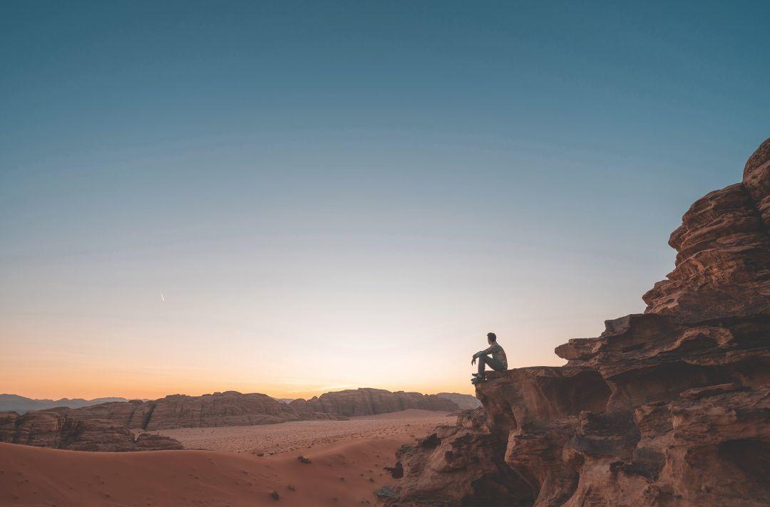 A photo of a man sitting on the edge of a rock, looking out over a desert canyon