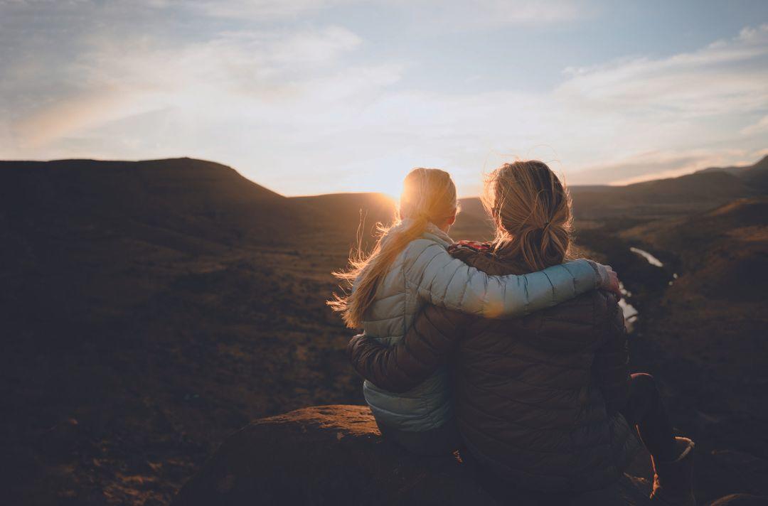 A photo of a mother and daughter with their arms around each other, sitting on a rock looking at the sun setting over hills