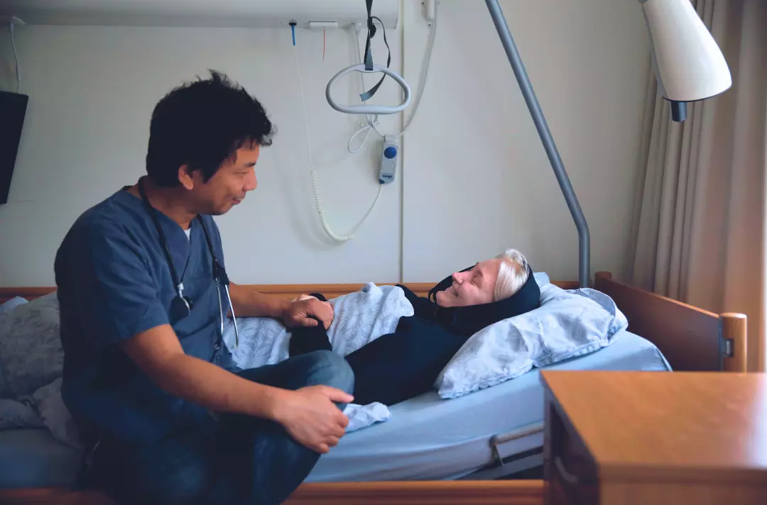 Photo shows someone lying in bed. Someone else sits on the bed. They are smiling broadly at each other.