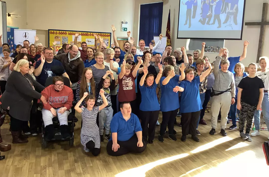 A photo shows a large group of members of the Music Man Project cheering at the camera.