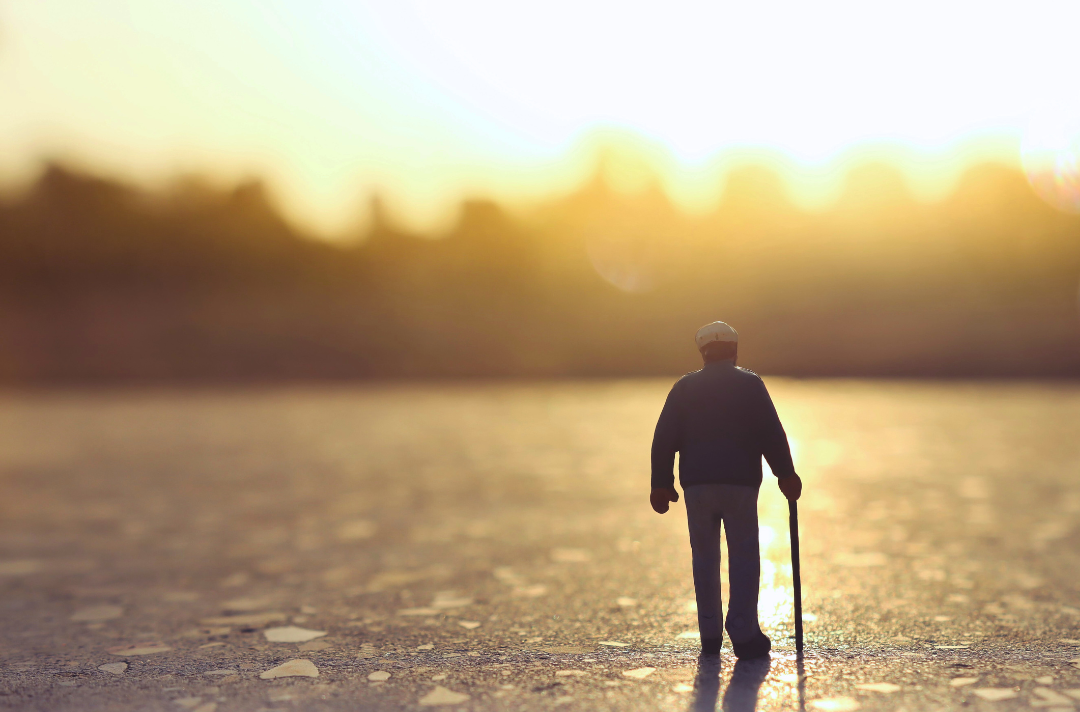 A photo shows a very small model representing an older man walking across a vast expanse towards the sunset and the horizon.