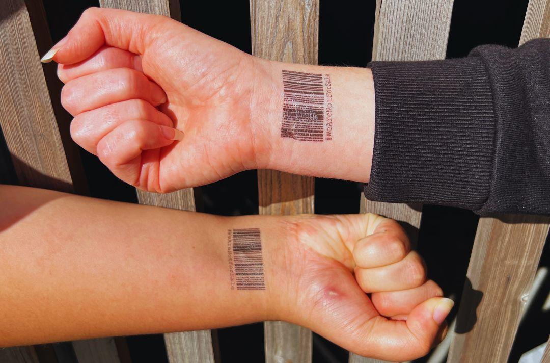 Two people clenching their fists with a barcode temporary tattoo on their wrists with the words We Are Not for Sale'