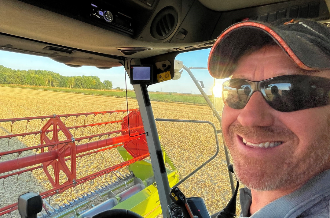 A selfie shows a man in a combine harvester smiling at the camera.