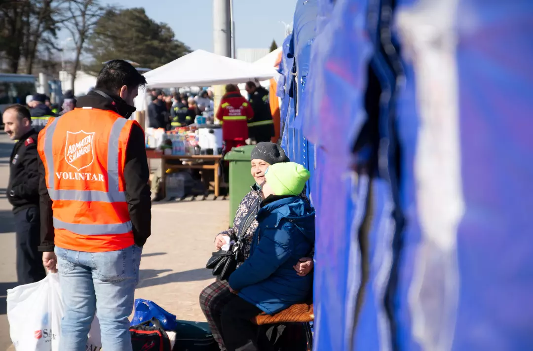 A photo shows someone in an orange Salvation Army branded hi viz vest talking to two people sat wrapped up in warm clothing.