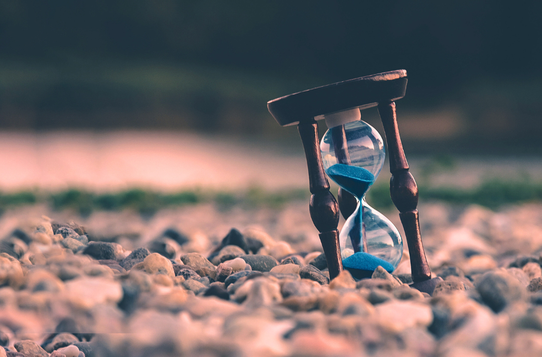 A photo shows an hourglass filled with blue sand sitting on a stony beach.