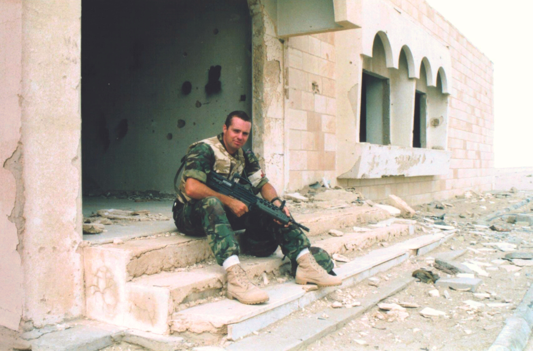 A photo shows Kevin Russell sat in military gear outside a building with debris scattered around.